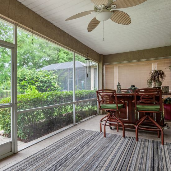 The interior of a screened-in porch with a striped rug and bar with two barstools inside on the right and a yard with bushes and trees outside of the screen wall and door on the left.